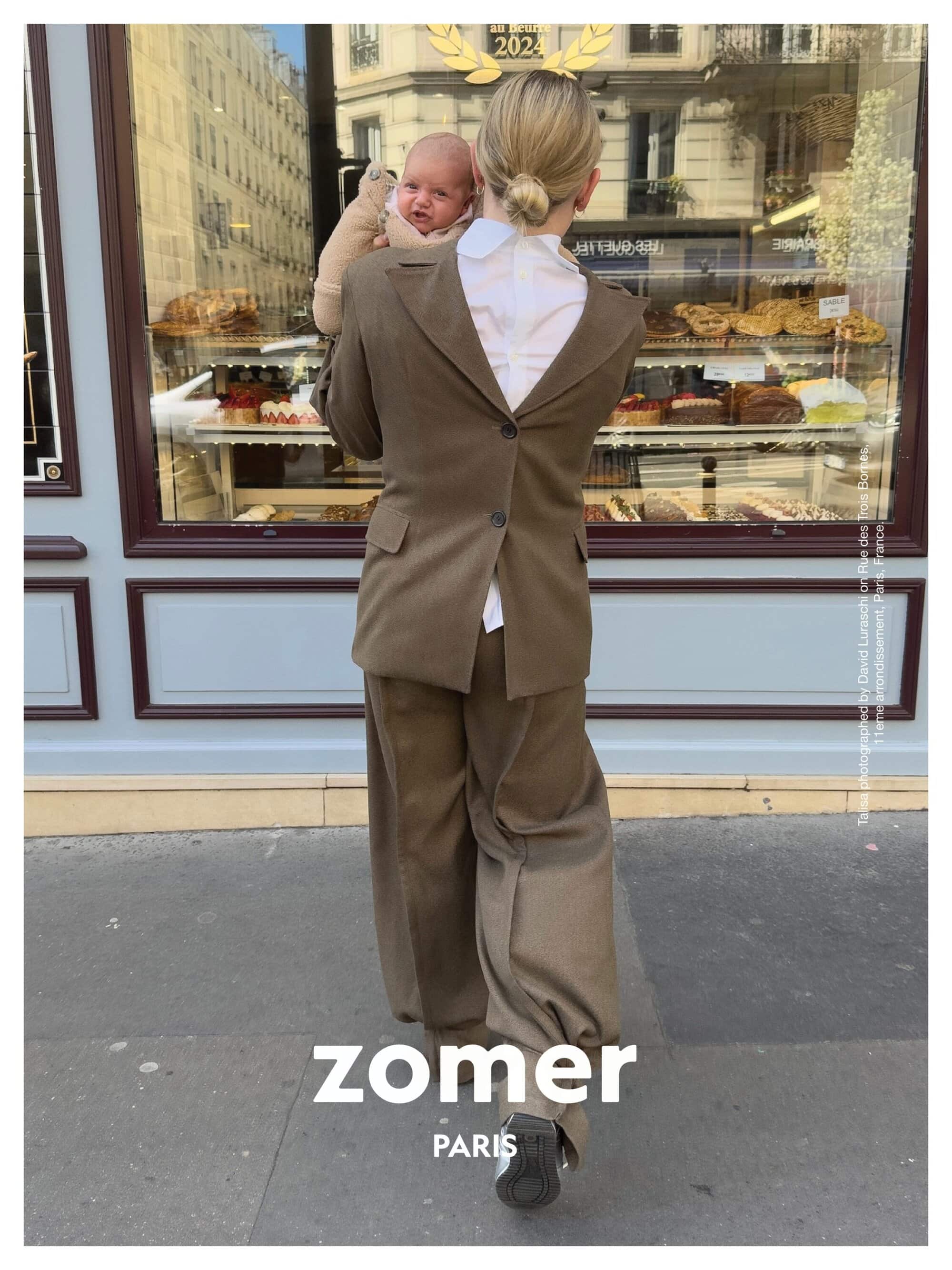 Woman wearing a backwards brown suit holding a baby in front of a Parisian bakery storefront