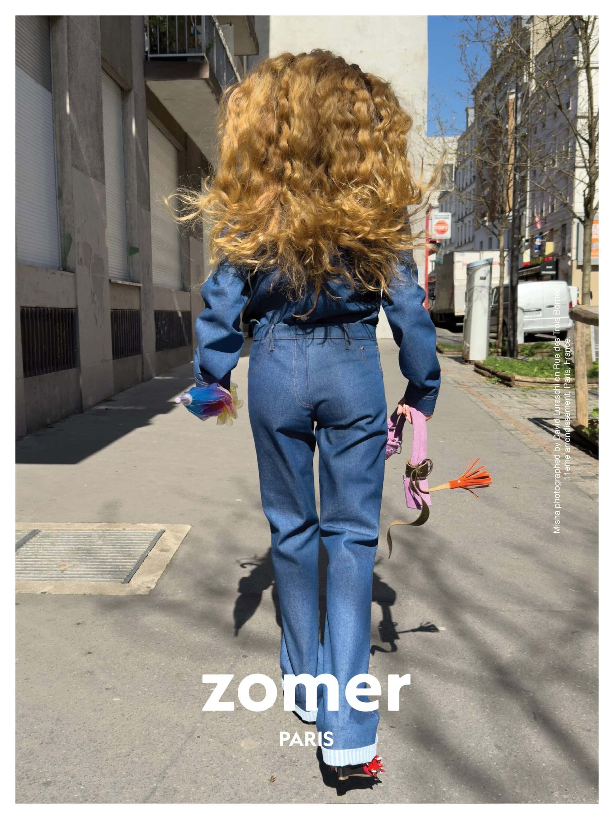 Model seen from behind walking down a city street wearing a blue denim jumpsuit with long blonde hair and holding a small handbag.