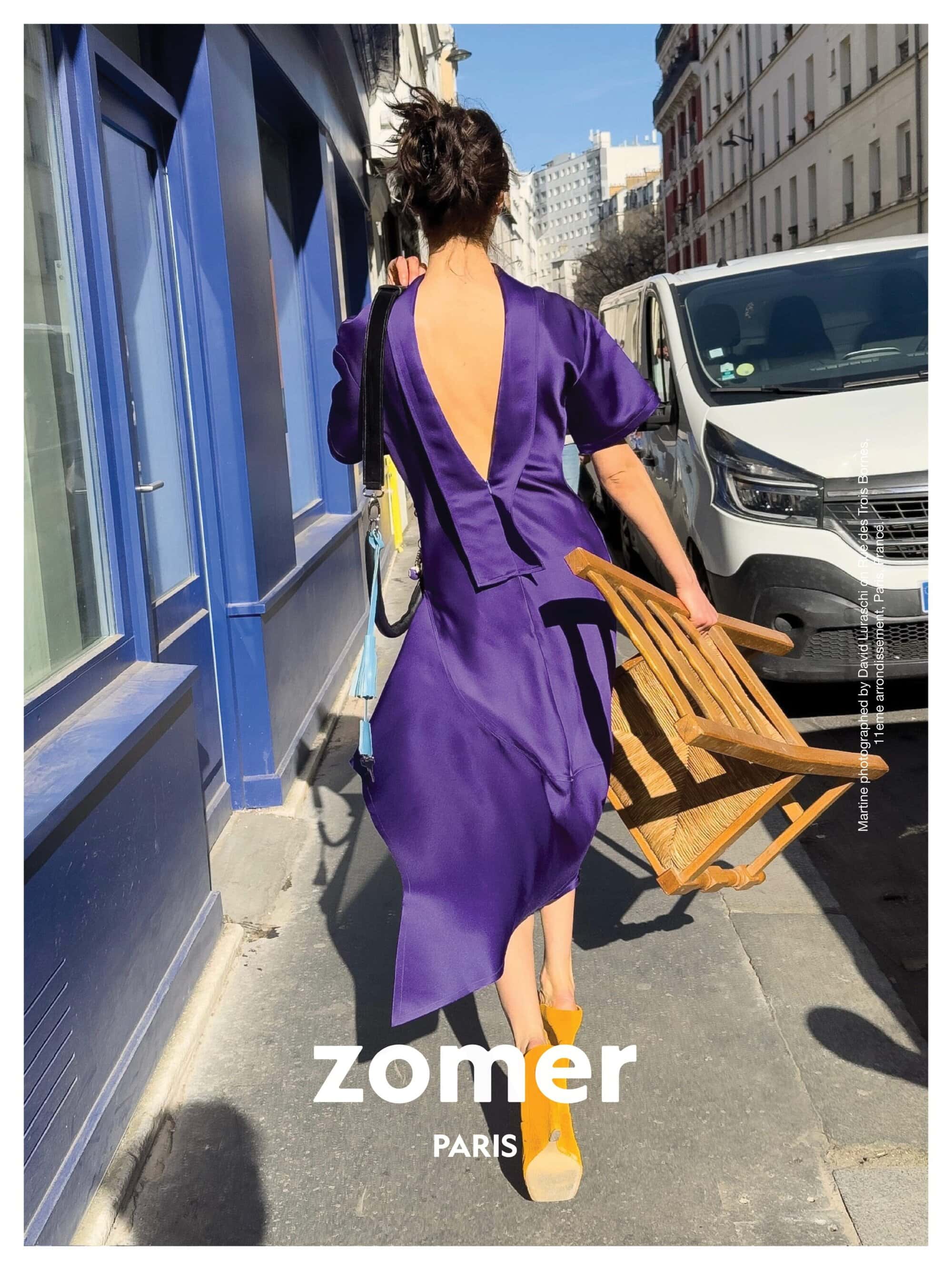 Model wearing a backless purple dress walking down a sunny city sidewalk holding a wooden chair
