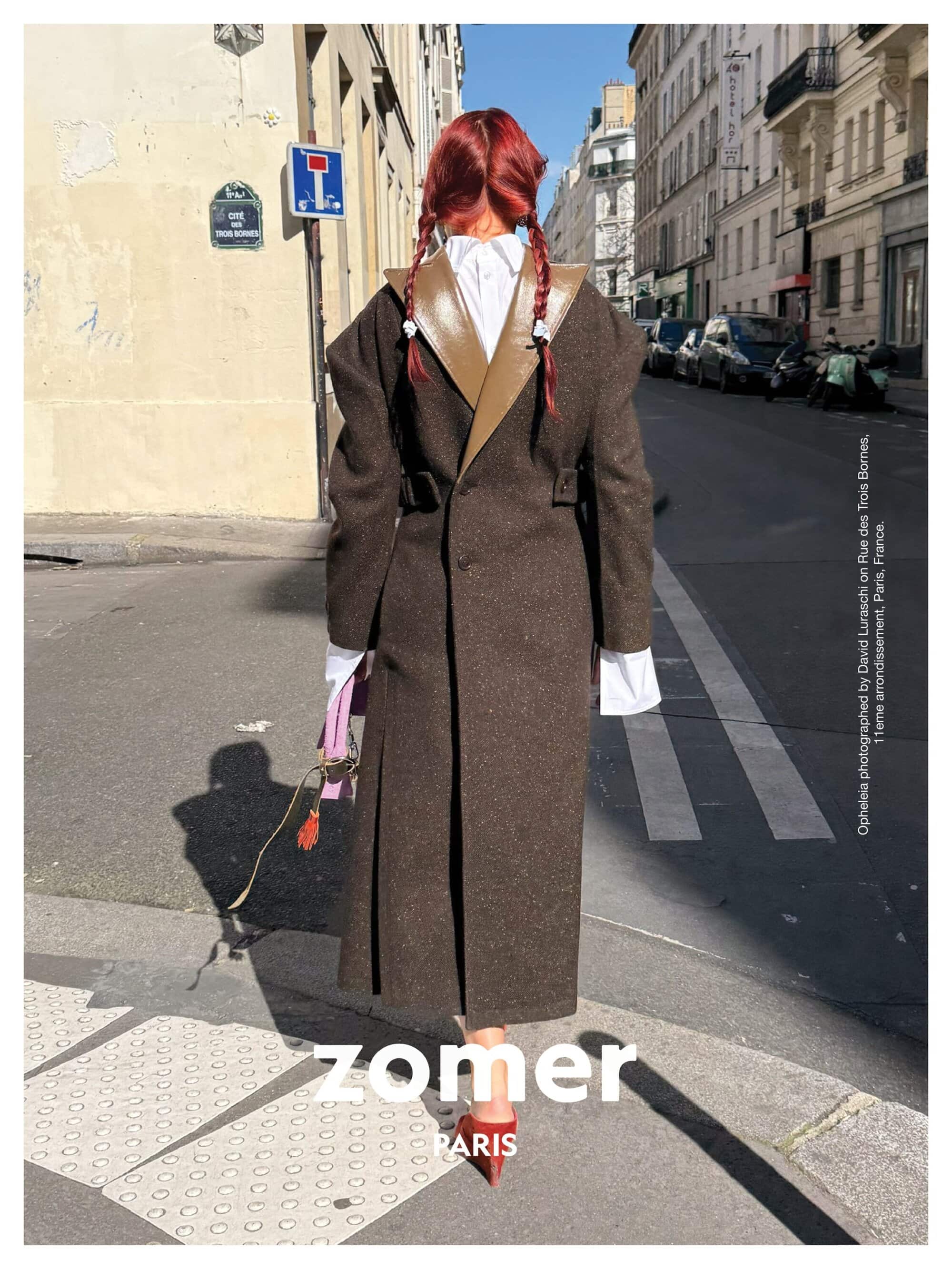 Model seen from behind with red braided hair wearing a long brown coat walking on a Parisian street