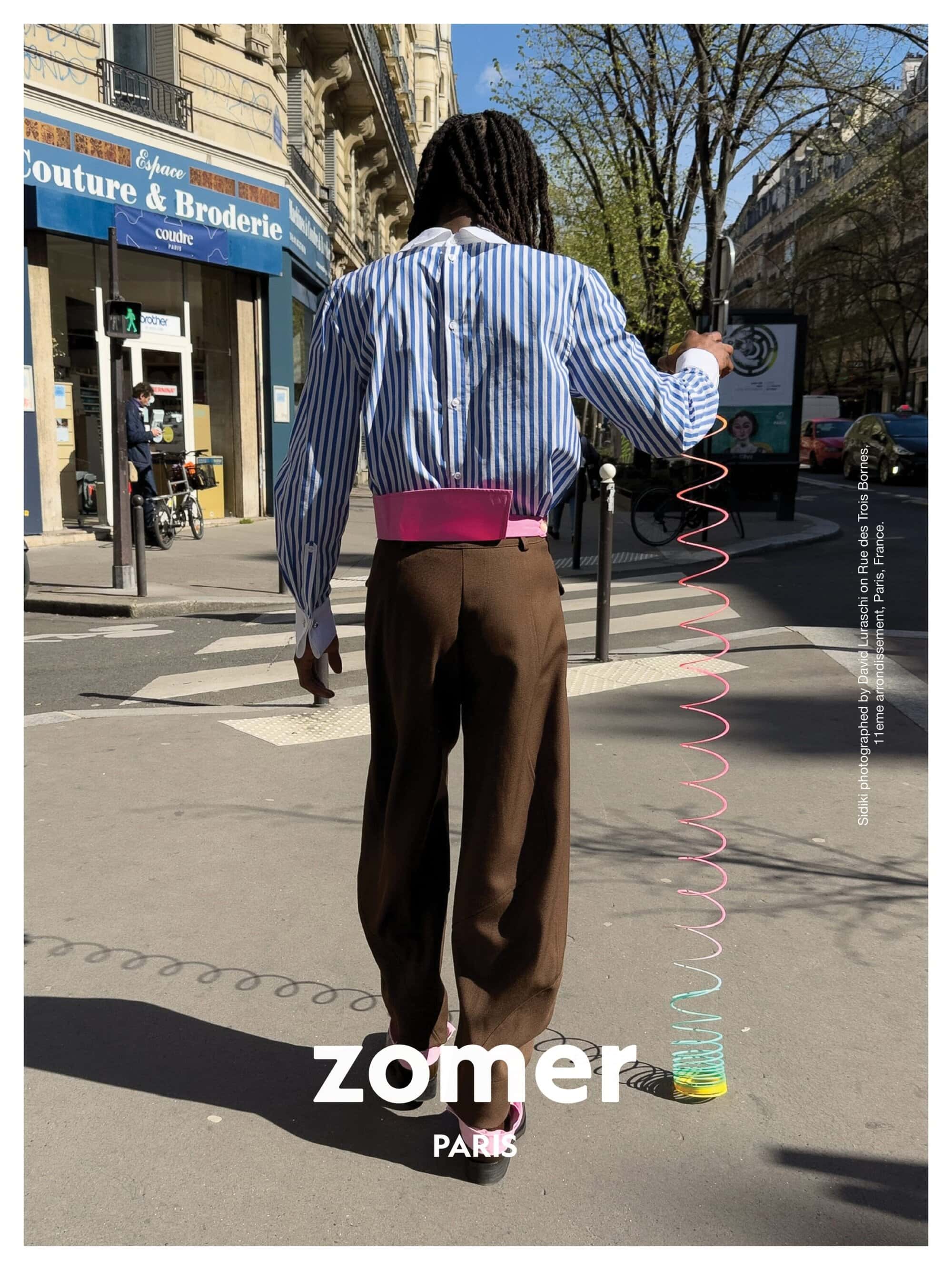 Model with dreadlocks wearing striped shirt and brown trousers walking on a sunny city street holding a colourful toy spring