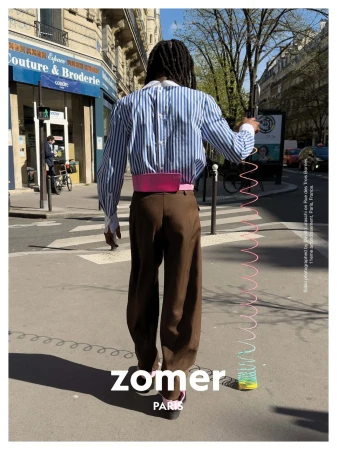 Model with dreadlocks wearing striped shirt and brown trousers walking on a sunny city street holding a colourful toy spring