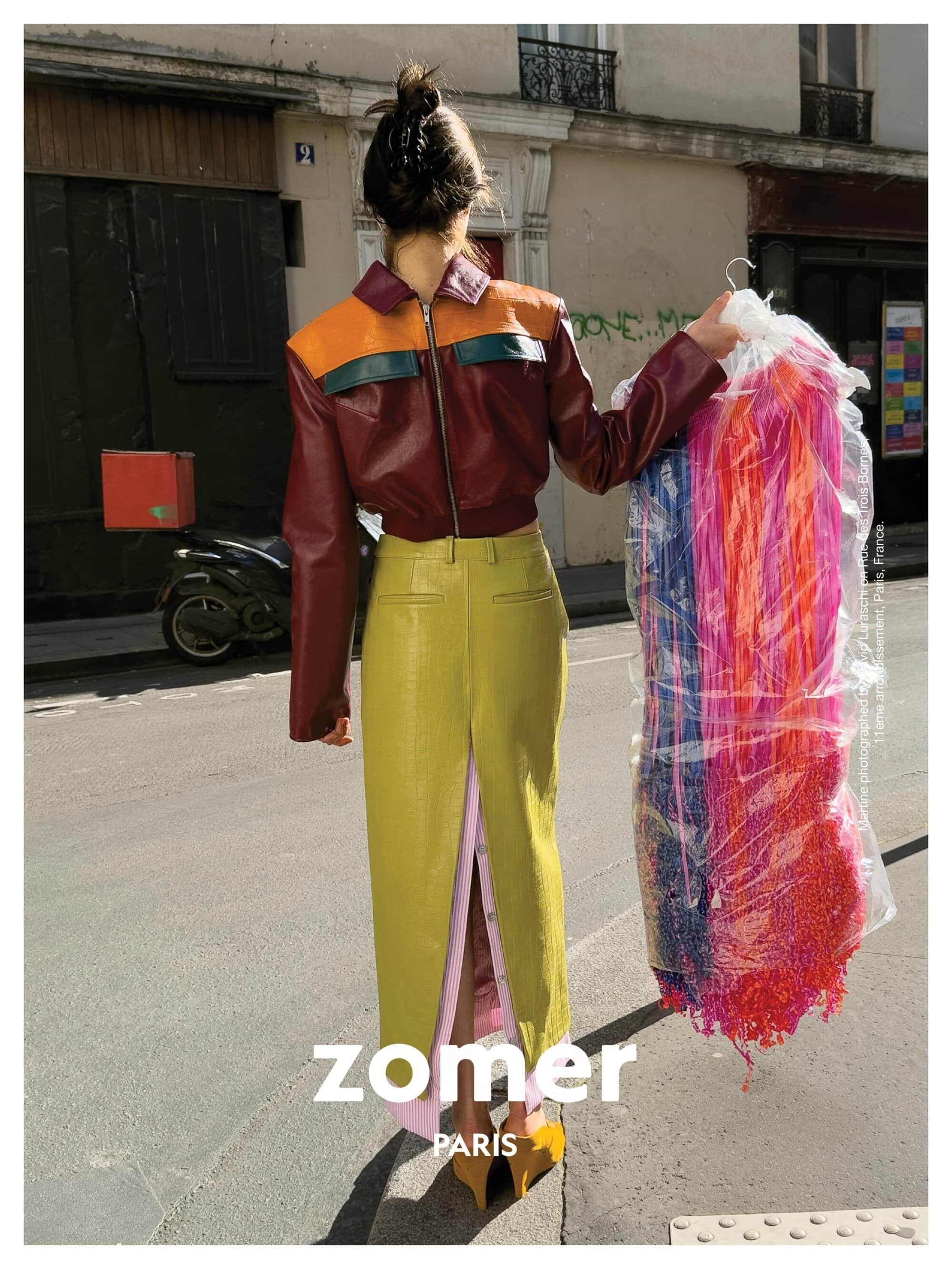 Model standing on a city street holding a garment bag, wearing a color-blocked leather jacket and yellow skirt.