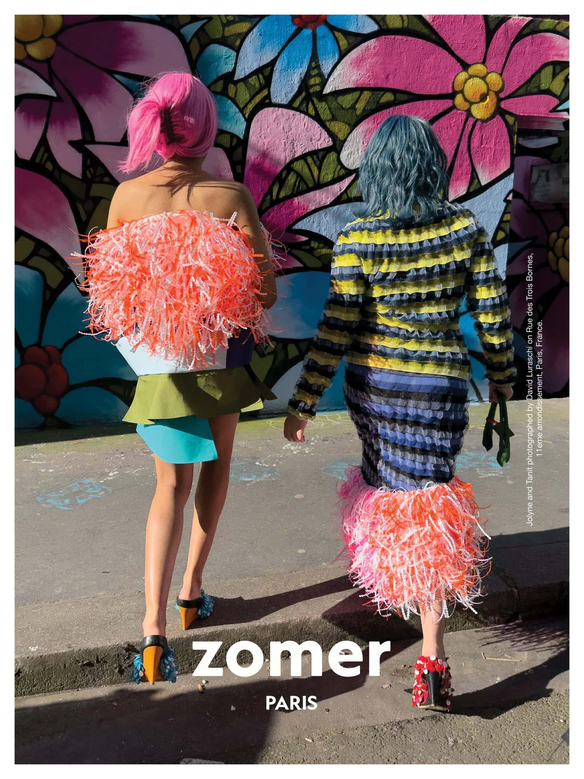Two models with bright pink and blue hair wearing textured colorful fashion pieces walking in front of a painted floral wall