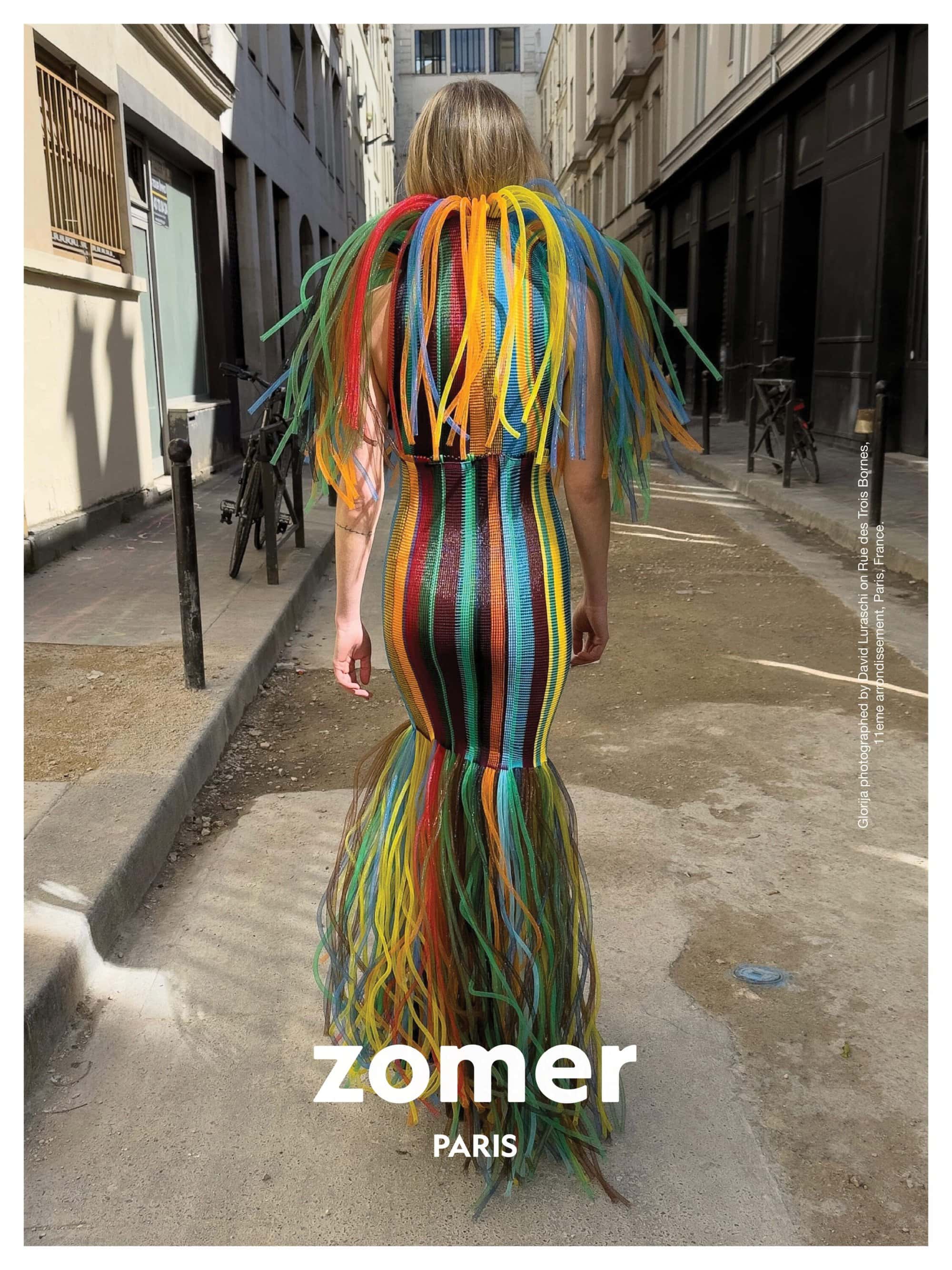 Model from behind wearing a brightly colored striped gown with fringed collar walking down a city alleyway