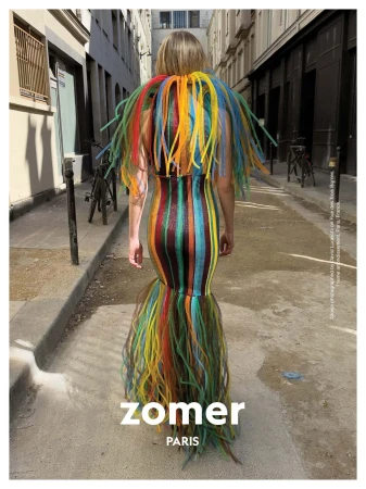 Model from behind wearing a brightly colored striped gown with fringed collar walking down a city alleyway