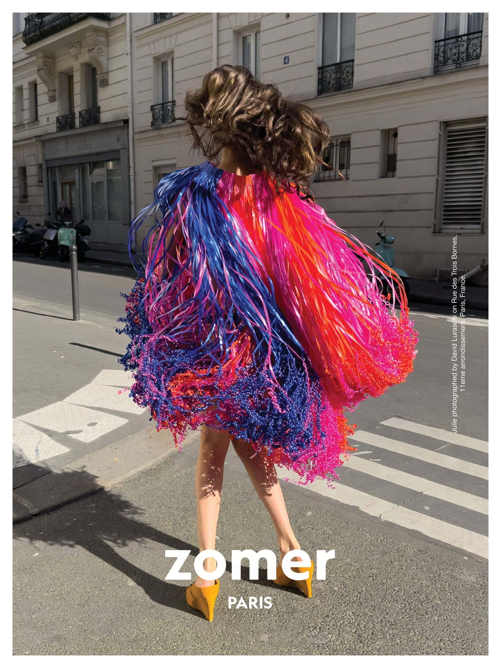 Model seen from behind wearing a vibrant multicolored fringe jacket and yellow wedge shoes on a sunny Parisian street
