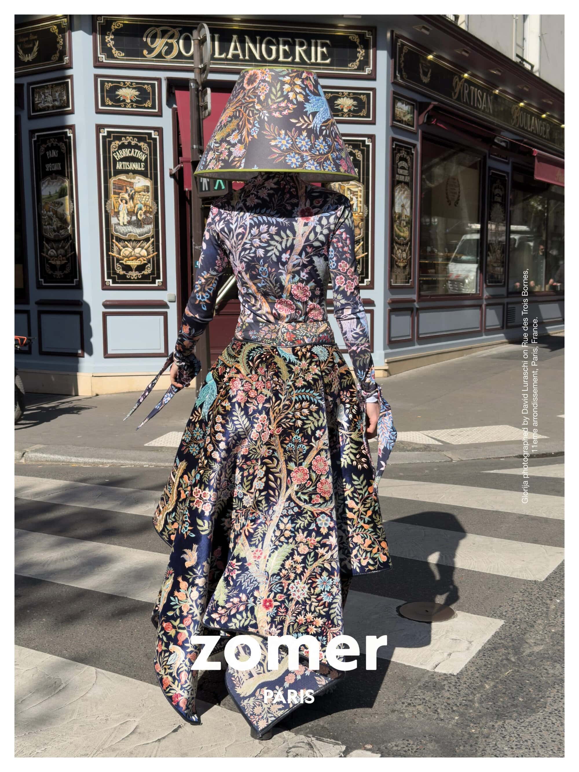 Model wearing a matching floral patterned outfit and lampshade headpiece standing on a street corner in front of a bakery.