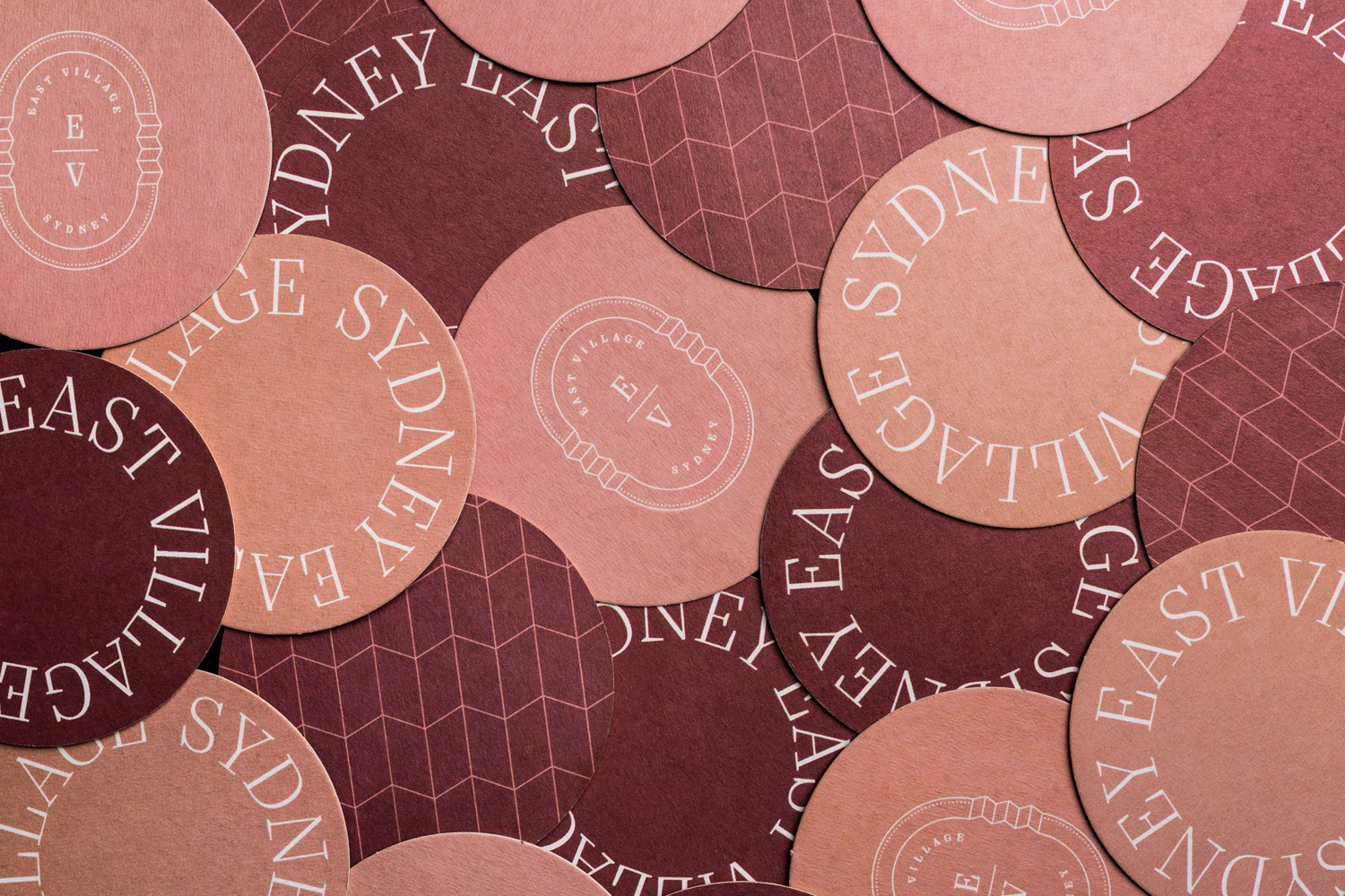Flat lay of overlapping round cardboard coasters featuring brand typography and geometric patterns in shades of burgundy and pink.