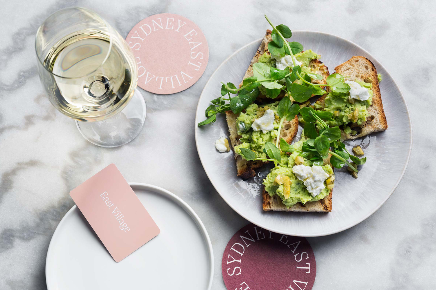 Avocado toast with greens on a grey plate, a glass of white wine, and branded pink coasters and card on a marble surface.