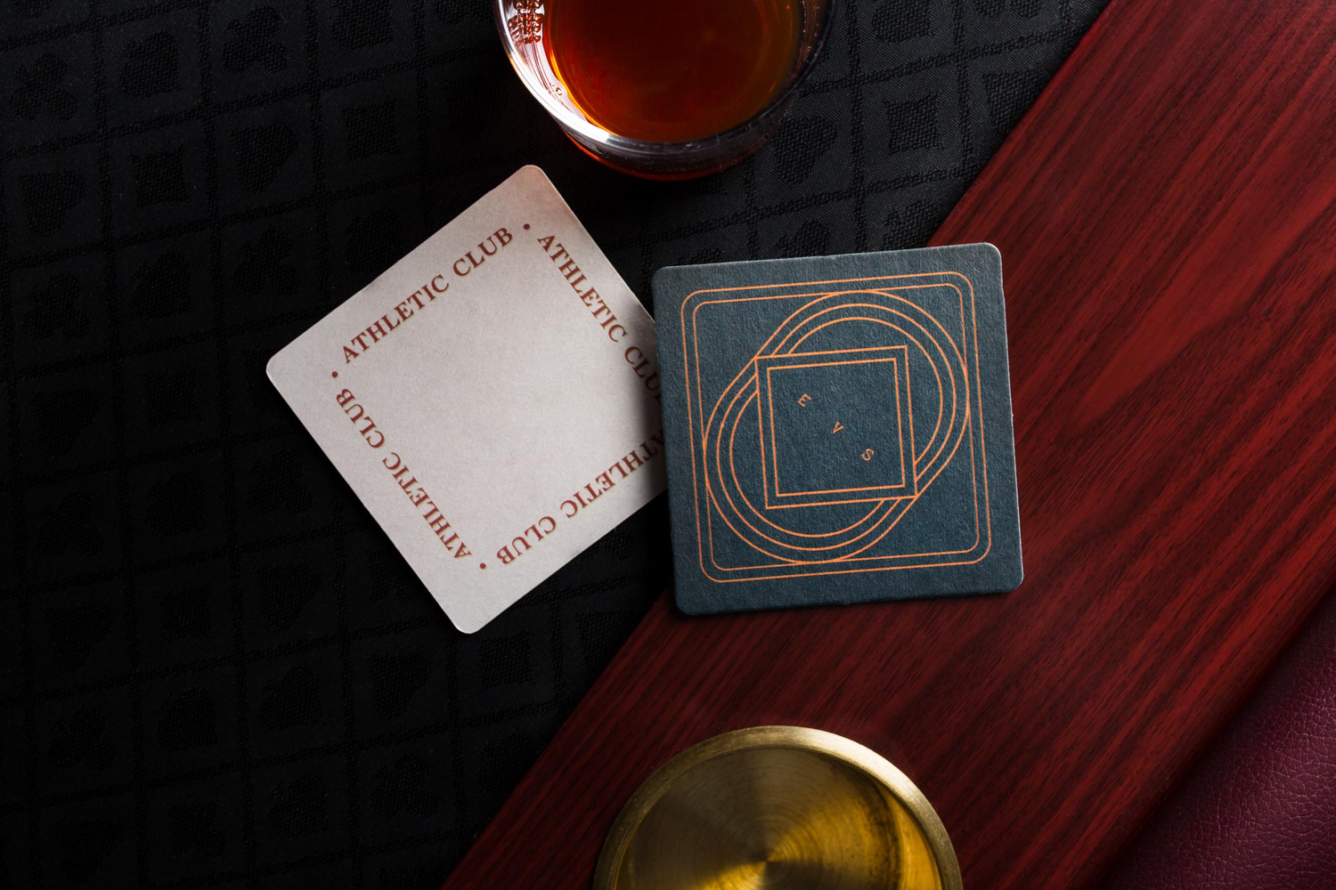 Top-down view of branded drink coasters, a glass of amber liquid, and a wooden tray on a dark textured surface.
