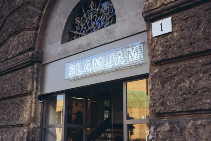 Storefront exterior with a glowing neon Slam Jam sign above glass doors, set in a rustic stone building facade.