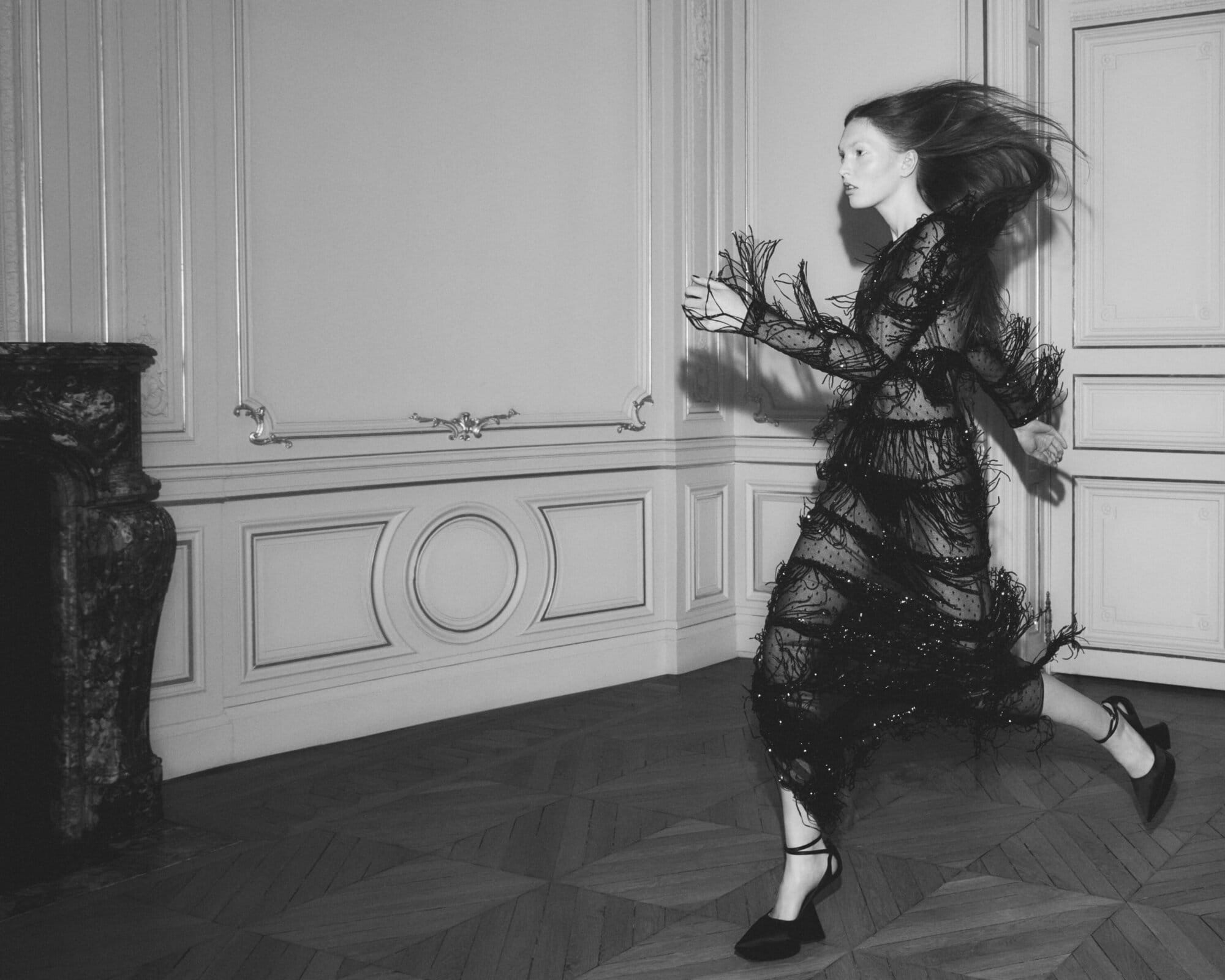 Model in black feathered sheer dress running across a classical room with wooden floors for Nina Ricci Fall 2025 campaign.