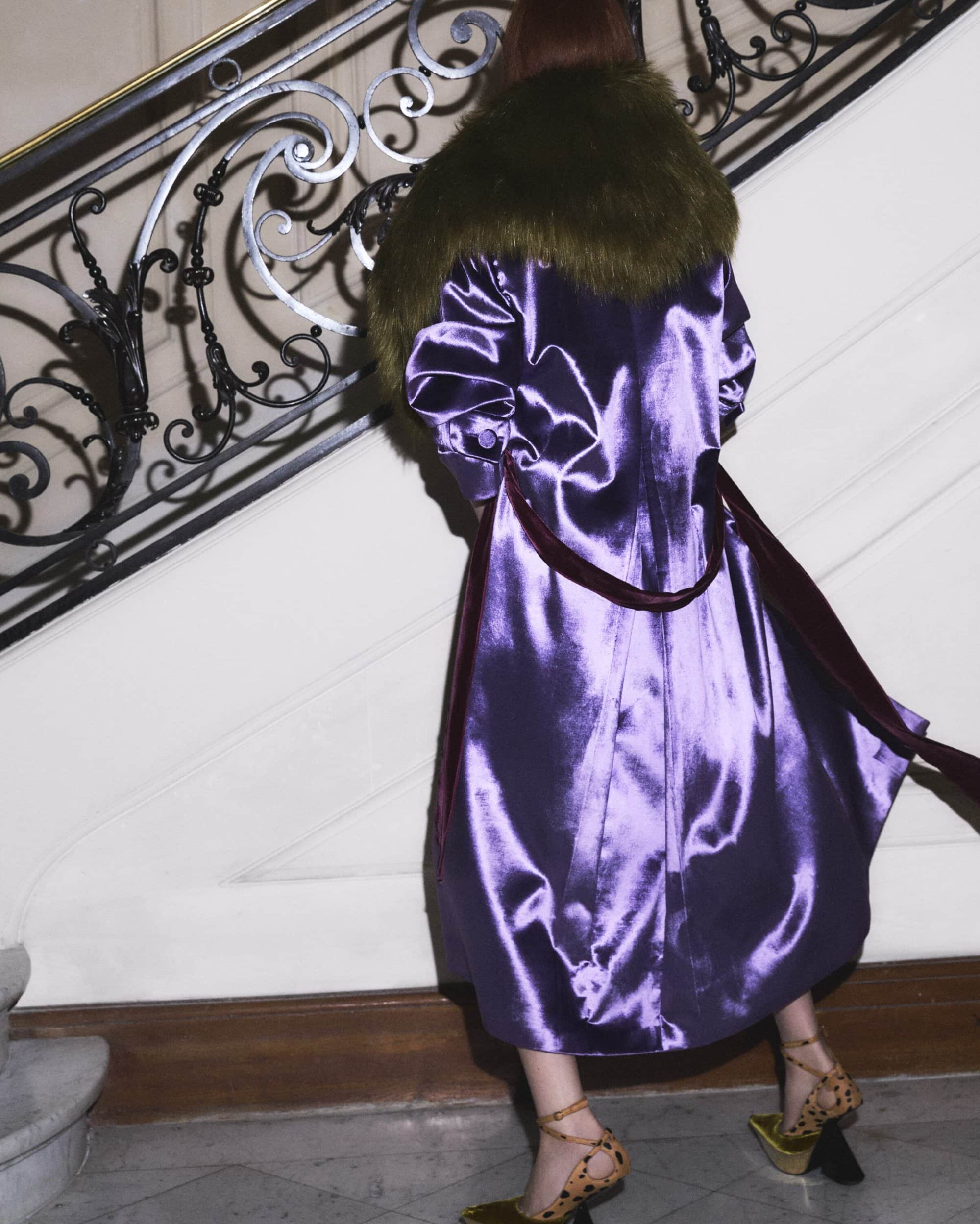 Model from behind in a glossy purple satin coat with a furry green collar standing on a stone staircase