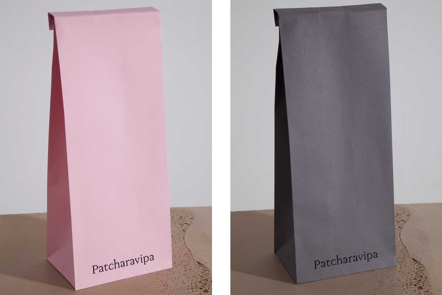 Two tall paper bags in light pink and dark grey with brand text standing on a textured neutral surface.