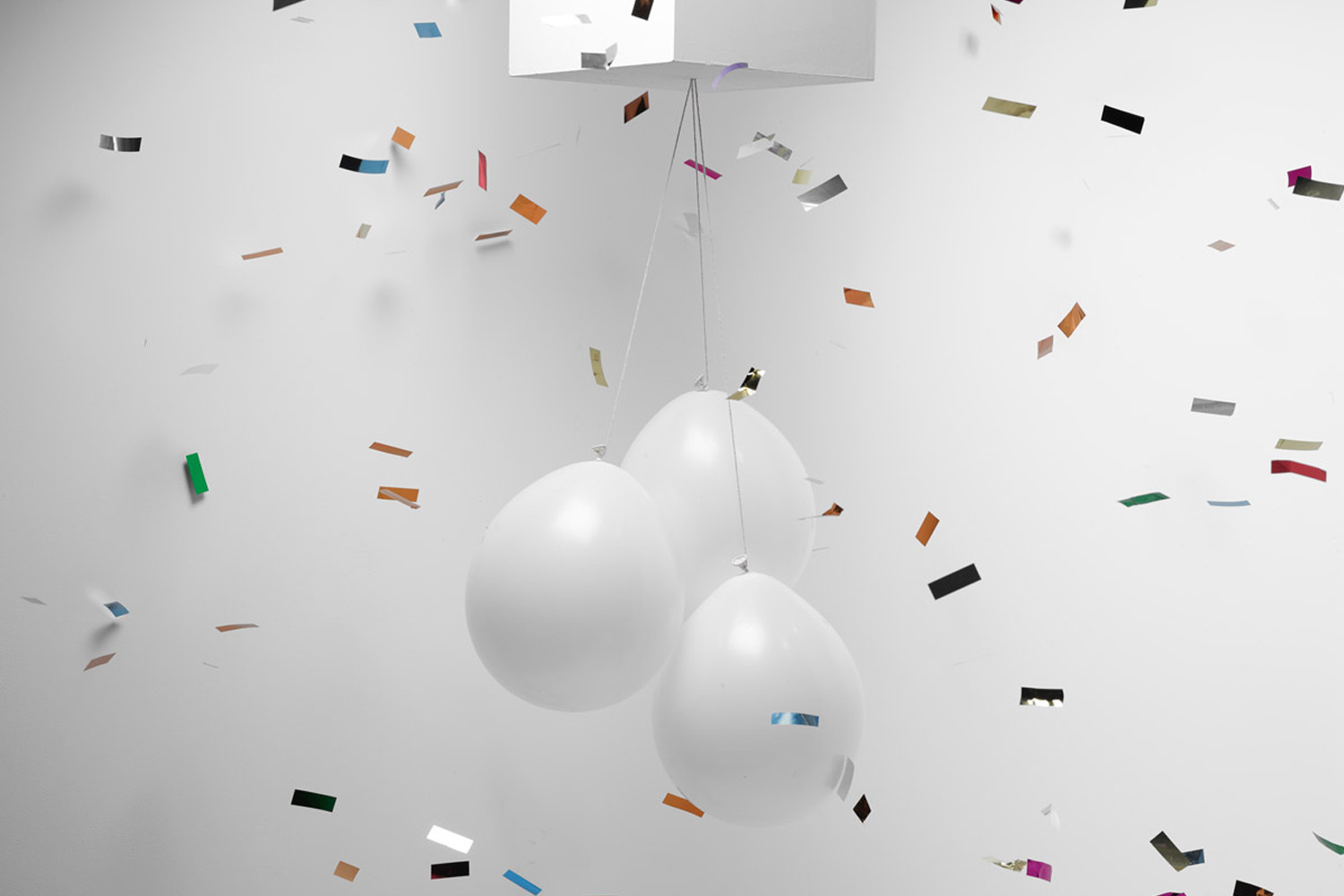 Three white balloons suspended by strings against a plain white background amid falling metallic confetti pieces.