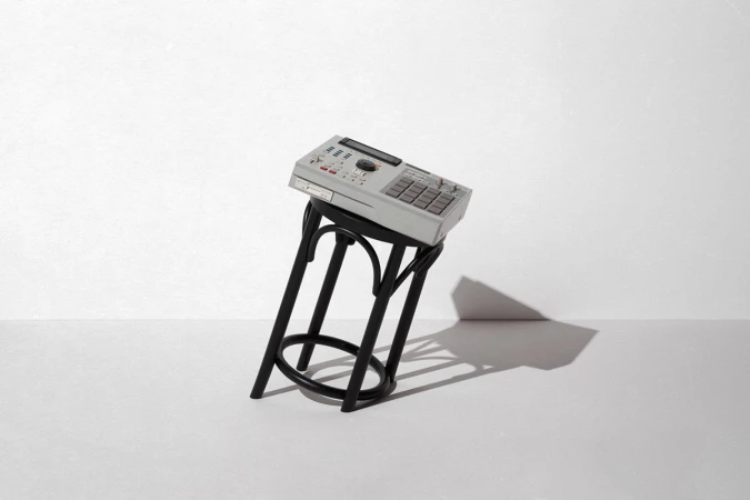 Vintage grey music sampler resting diagonally on a black wooden stool against a neutral white wall.