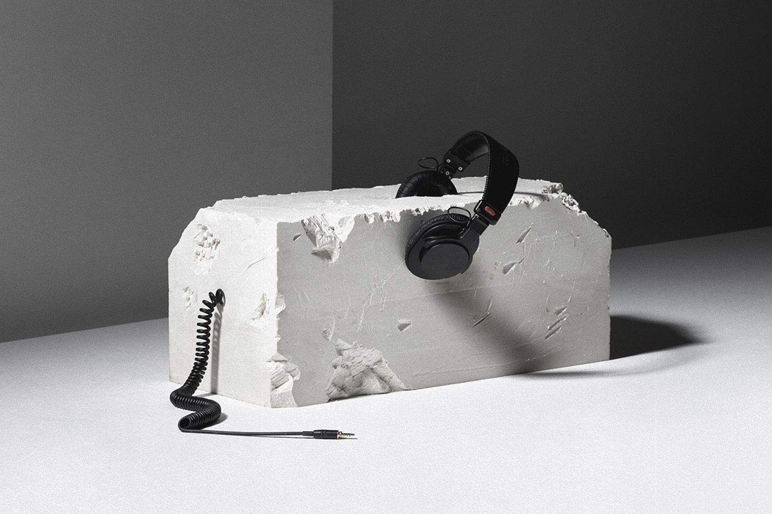 Black over-ear headphones resting on a textured white rectangular stone block with a coiled audio cable extending out.