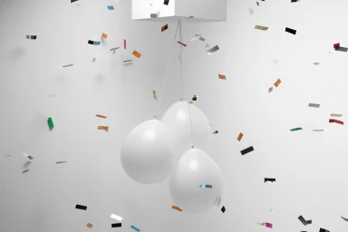 Three white balloons suspended by strings against a plain white background amid falling metallic confetti pieces.