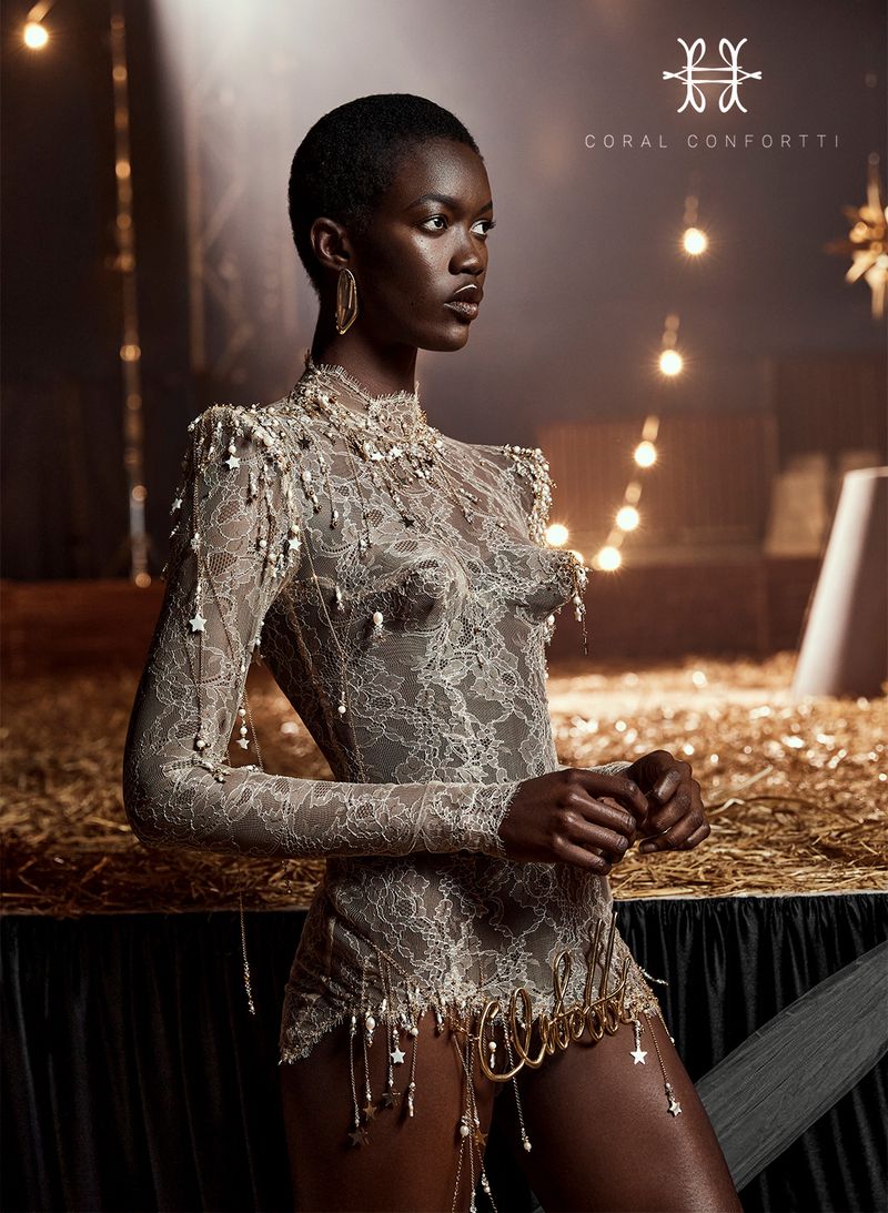 Portrait of a dark-skinned woman wearing a lace bodysuit with beaded embellishments in a warm, indoor setting with string lights.