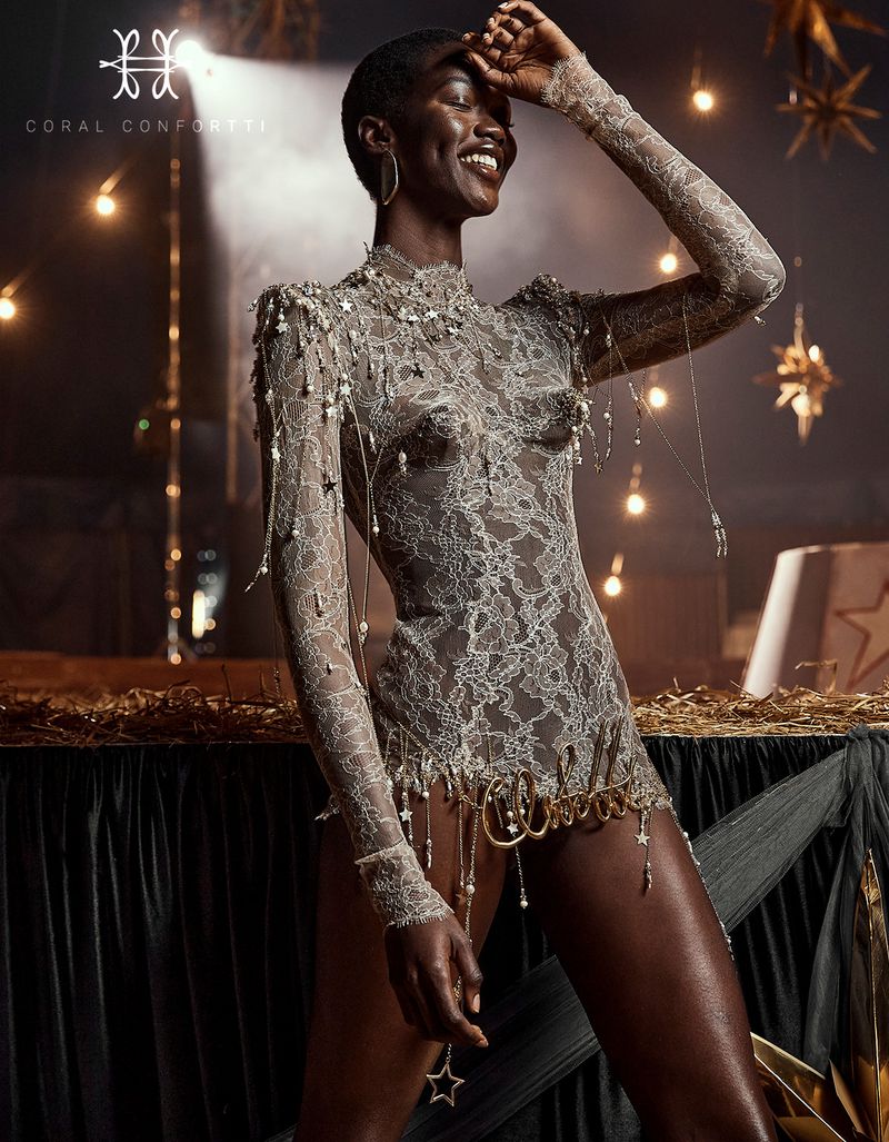 Stunning portrait of a Black woman in a silver lace bodysuit with jewelry. She is posing in a warm, festive indoor setting.