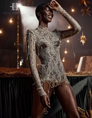 Stunning portrait of a Black woman in a silver lace bodysuit with jewelry. She is posing in a warm, festive indoor setting.