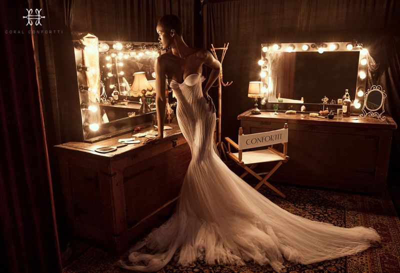 Woman in elegant white gown poses in vintage dressing room. Two vanities with lighted mirrors flank the model with dark curtains.