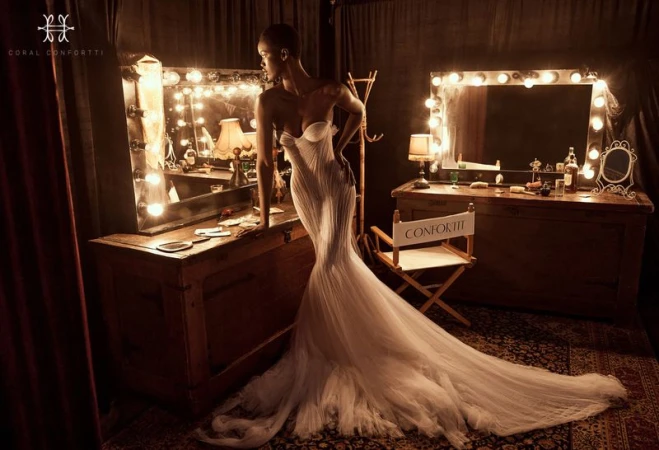 Woman in elegant white gown poses in vintage dressing room. Two vanities with lighted mirrors flank the model with dark curtains.