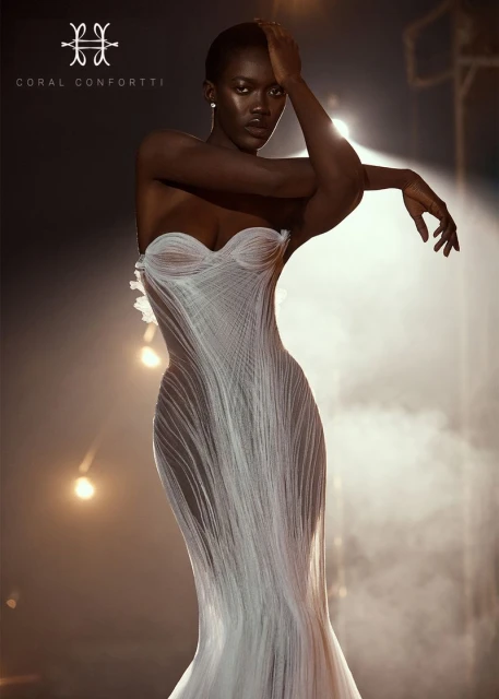 Elegant Black woman poses in strapless white gown. Studio lighting highlights the gown's layered, textural design with a dark, dramatic backdrop.