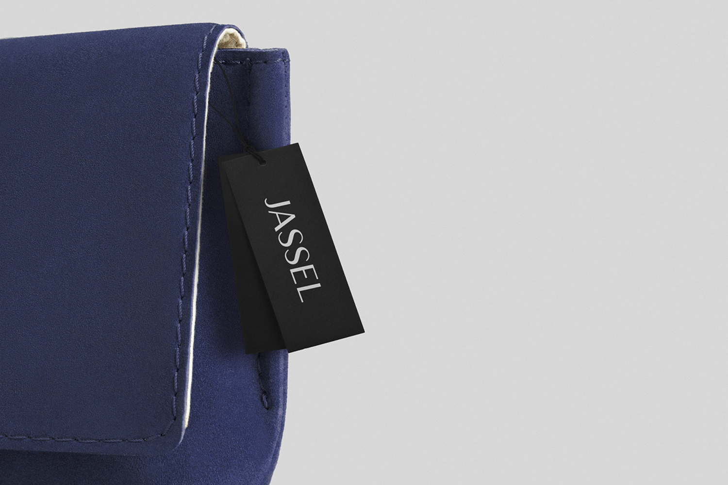 Close up of a dark blue suede handbag with visible stitching and a black brand tag reading Jassel against a plain grey background