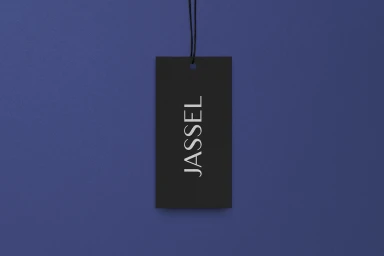 Black clothing tag with white text reading Jassel suspended by a black string against a textured blue background