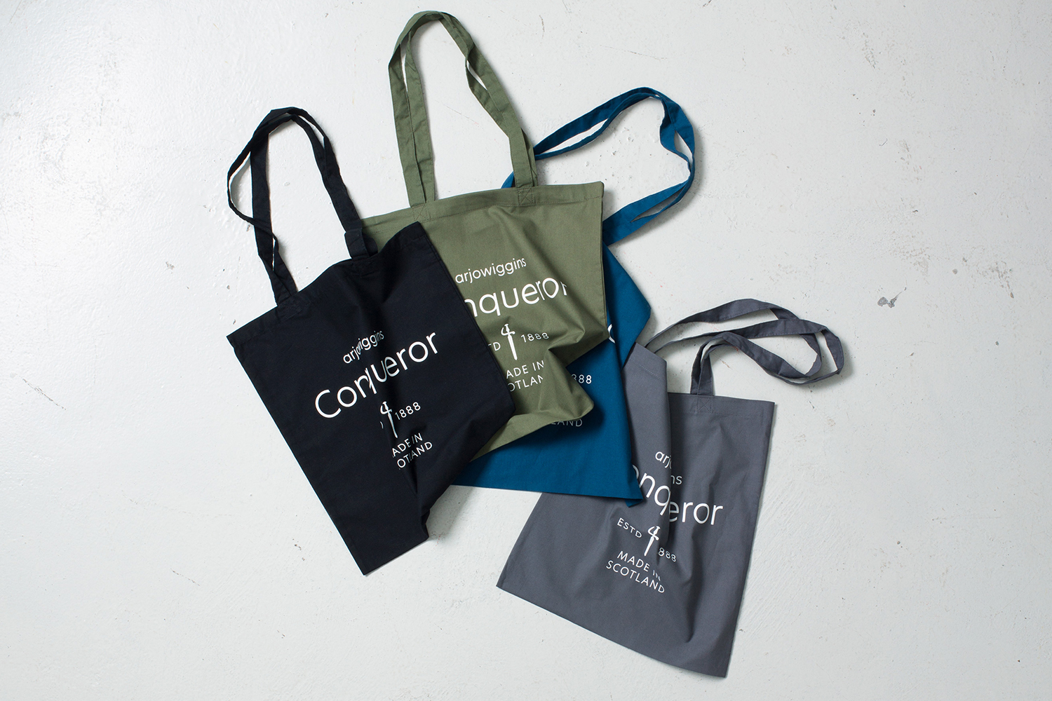 Four canvas tote bags in black, olive green, blue, and grey arranged on a white seamless background featuring branded typography.