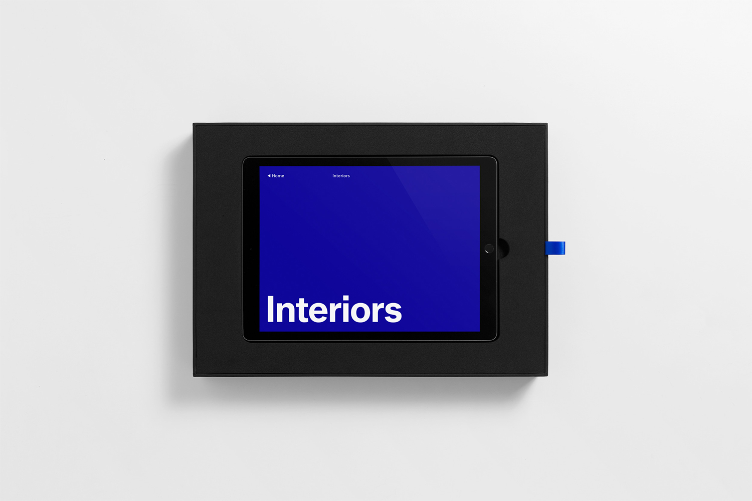 A tablet device displaying a blue screen with the word Interiors resting inside a black presentation box on a white seamless background