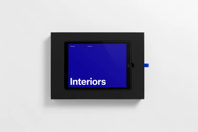 A tablet device displaying a blue screen with the word Interiors resting inside a black presentation box on a white seamless background