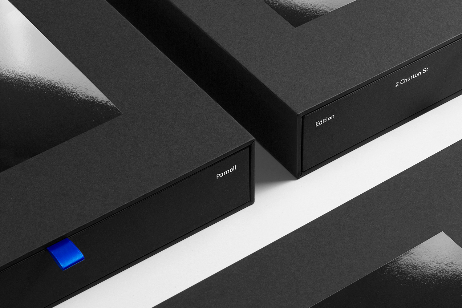 Close-up of black textured packaging boxes with typography and a bright blue ribbon pull tab on white surface