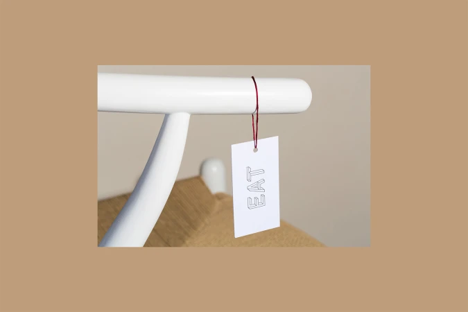 White paper tag reading Eat hanging by a red string from a white wooden chair rail against a plain background