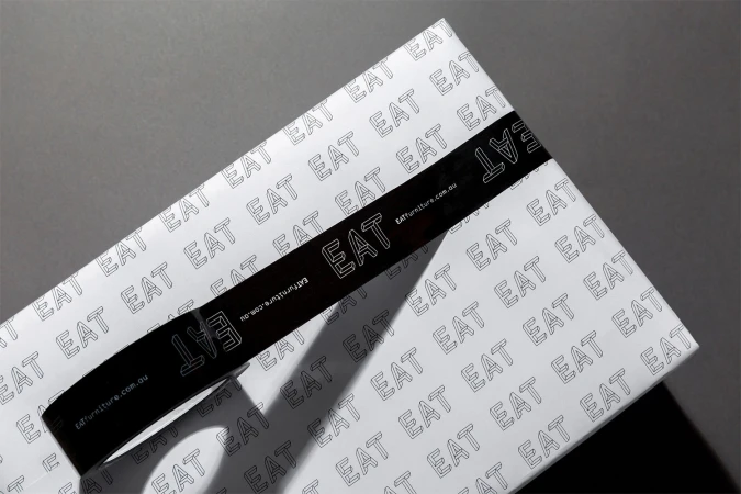 White packaging box with repeating Eat logo pattern sealed with glossy black branded tape on a dark surface.
