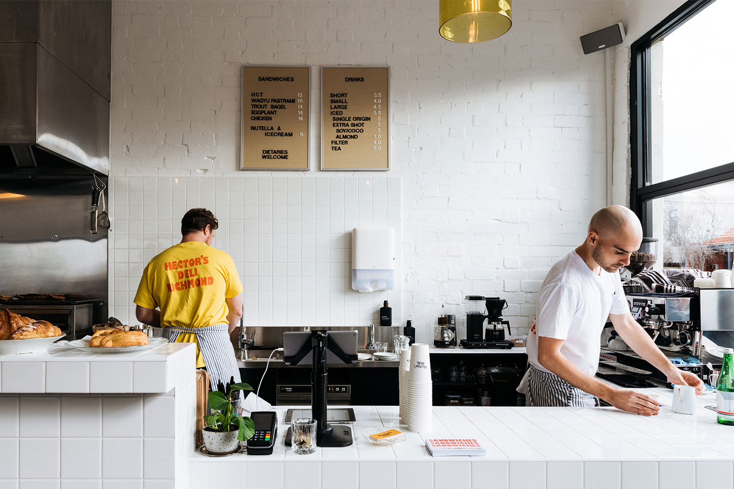 Two staff members working behind the counter inside Hector's Deli with white tiled walls and menu boards
