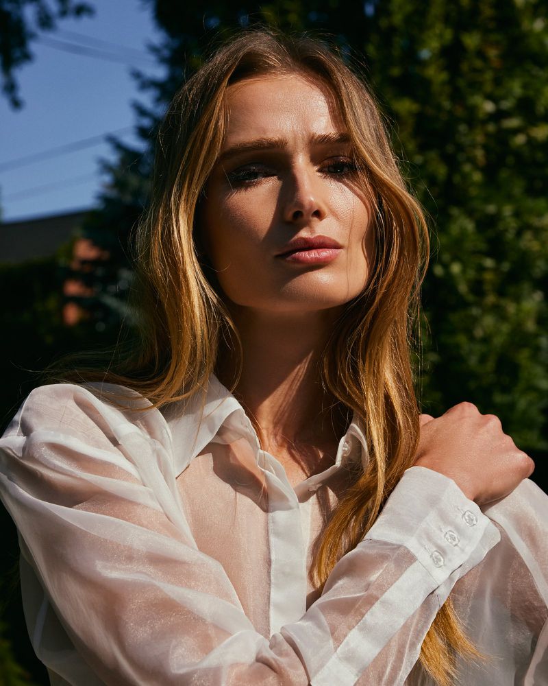 Close-up portrait of a blonde woman in a sheer white blouse, posing outdoors with trees and a blue sky visible in the background.