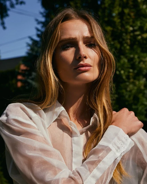 Close-up portrait of a blonde woman in a sheer white blouse, posing outdoors with trees and a blue sky visible in the background.