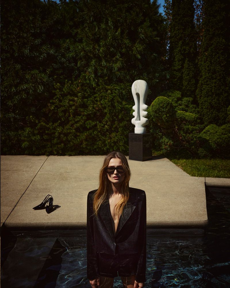 A woman in a black blazer stands in a pool with high heels and a modern sculpture nearby, set against a backdrop of green trees.
