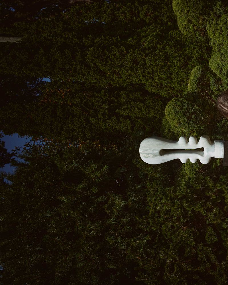 Abstract white sculpture of a guitar-like figure with rounded edges stands against a dark green background of dense, overgrown bushes.