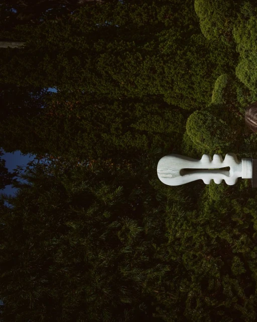 Abstract white sculpture of a guitar-like figure with rounded edges stands against a dark green background of dense, overgrown bushes.