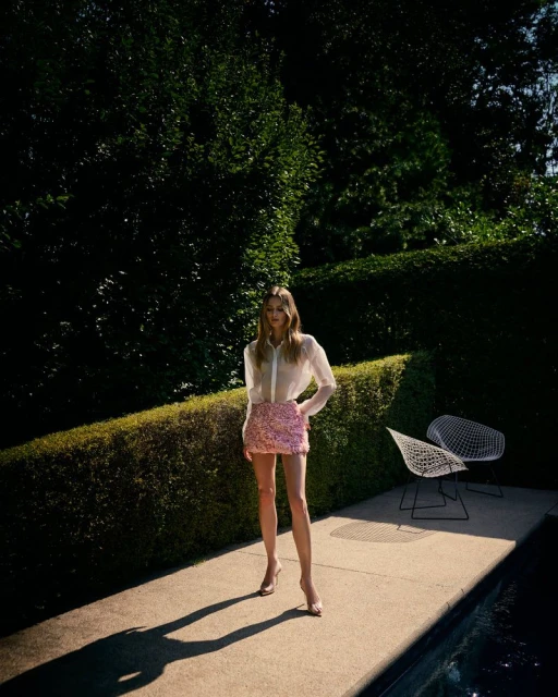 A full shot of a slender woman in a sheer white top and pink skirt standing on a concrete patio with a green foliage background.