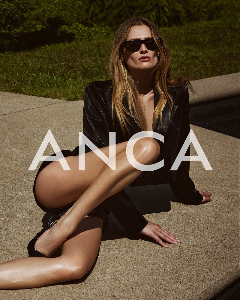 A woman in a black jacket poses outdoors, sitting on a concrete surface, wearing sunglasses, with green foliage in the background.