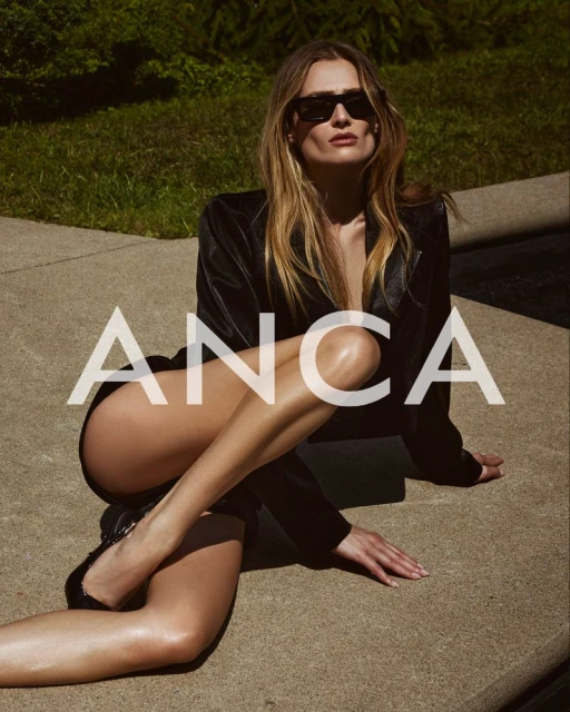 A woman in a black jacket poses outdoors, sitting on a concrete surface, wearing sunglasses, with green foliage in the background.