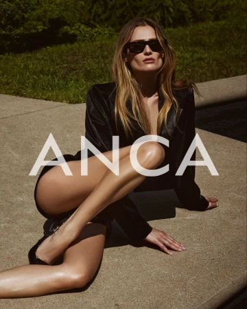 A woman in a black jacket poses outdoors, sitting on a concrete surface, wearing sunglasses, with green foliage in the background.