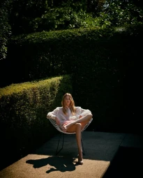 Model Anastasiia Koval seated in a wire chair outdoors wearing a white blouse against a dark green hedge background.