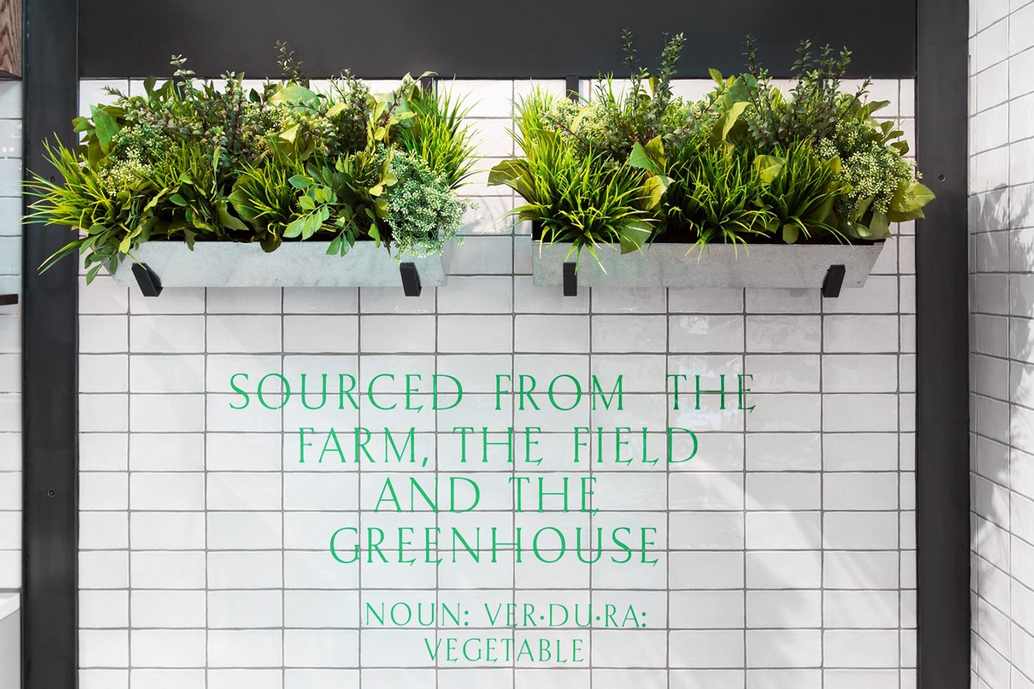White tiled wall with green text reading SOURCED FROM THE FARM, THE FIELD AND THE GREENHOUSE below two rectangular hanging planters