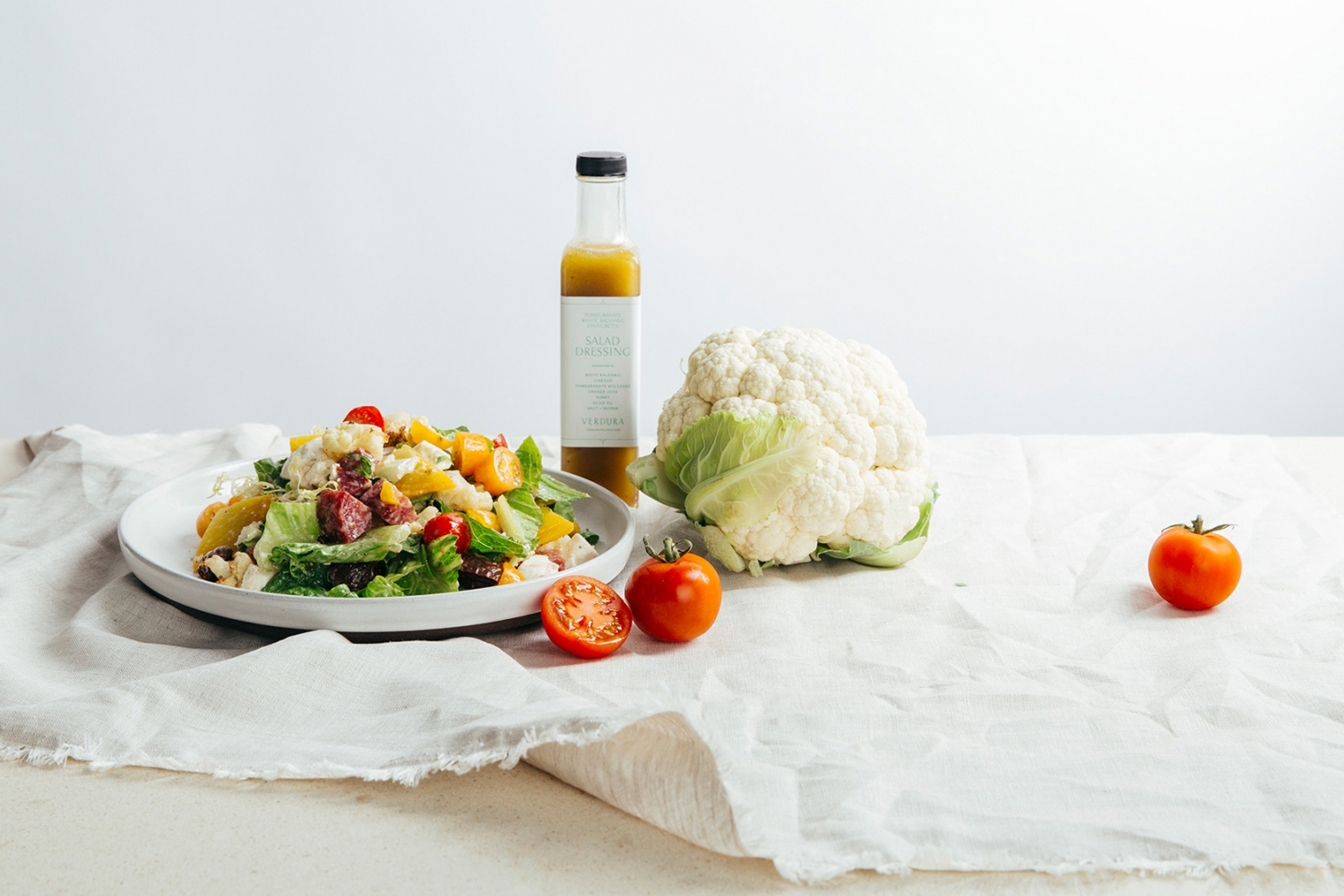 A white plate of chopped salad, a bottle of salad dressing, a whole cauliflower, and fresh tomatoes on linen fabric.